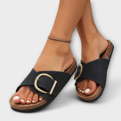 Zwart geklede vrouw draagt Avelina sandalen in beige, voeten gekruist.